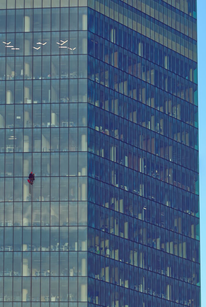A lone window cleaner in red scales a modern glass skyscraper, highlighting urban architecture.